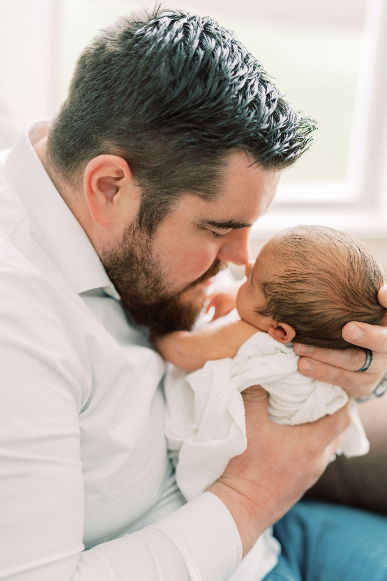 Bearded father in white shirt tenderly kissing his newborn baby wrapped in white blanket.