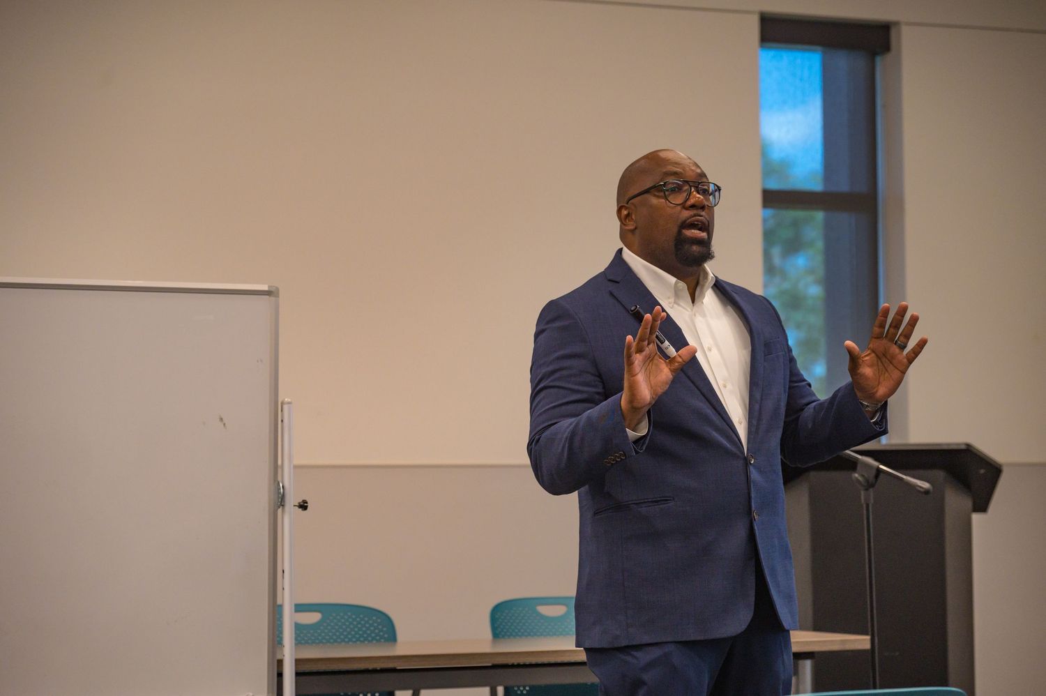 Man in blue suit presenting with both hands raised near a podium and whiteboard.