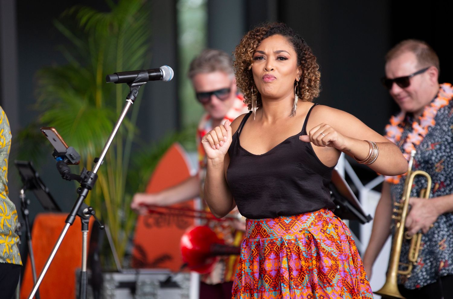 Female singer performing energetically on stage with band members playing trumpet and guitar at outdoor music event.