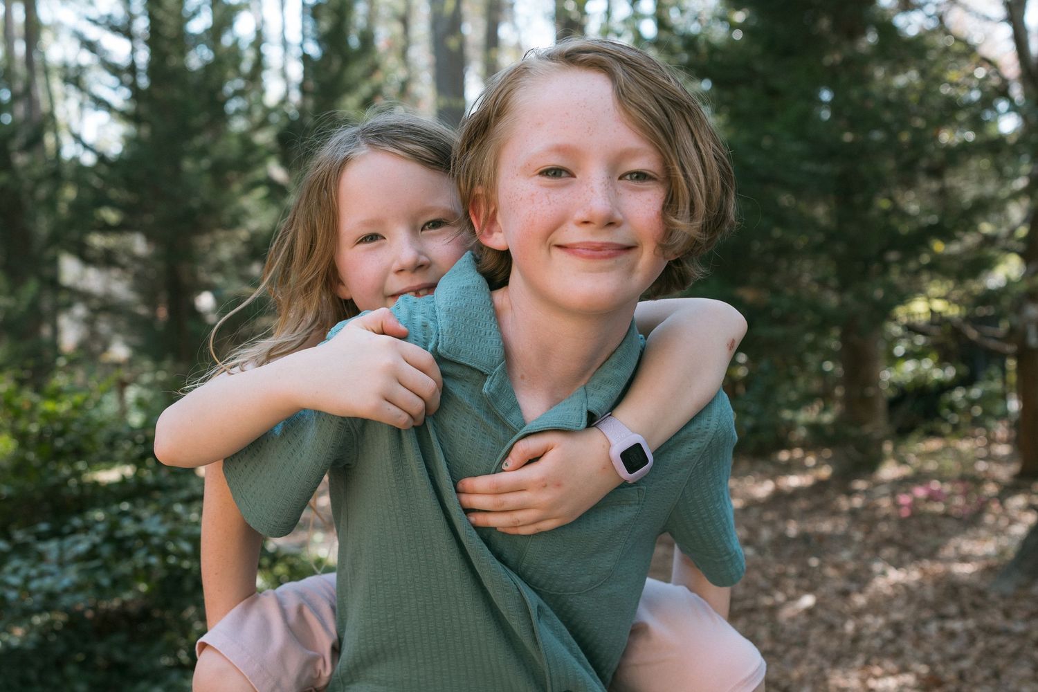Two smiling children share a piggyback ride outdoors in a lush green forest setting.
