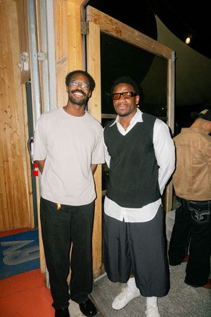 Two men smiling and posing together outdoors at night near a wooden door frame.