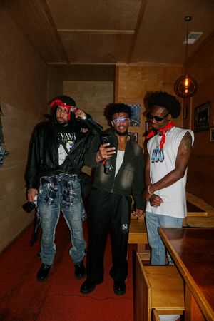 Three young men posing for a selfie indoors, wearing streetwear outfits with red accessories and stylish denim.