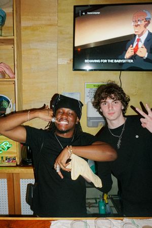 Two young men posing and smiling in a shop with a TV screen displaying "Behaving for the Babysitter" behind them.