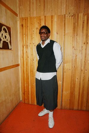 Man in black vest, white shirt, dark shorts, and white sneakers standing against a wooden wall.