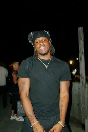 Young man in black t-shirt and bandana with gold chain and tattoos posing outdoors at night.