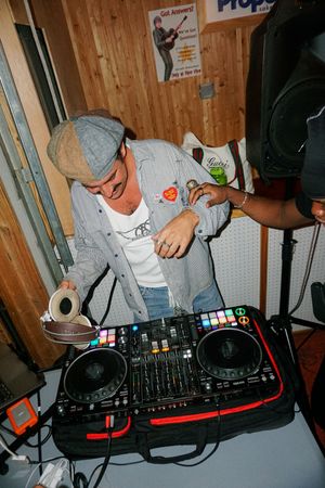 DJ mixing tracks on a Pioneer DJ controller at an indoor event, wearing a gray cap and denim shirt.