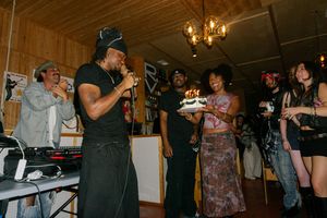 Group of people celebrating a birthday party indoors, with a man speaking into a microphone and a woman holding a cake.