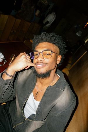 Young man with glasses and cross necklace smiling confidently at camera in dimly lit venue.