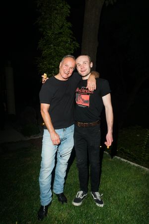 Two men in black t-shirts smiling and posing together outdoors at night on green grass.