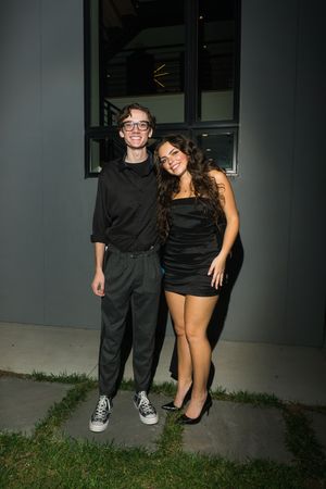 Young couple in all-black outfits posing outdoors at night, man in glasses and woman in mini dress.