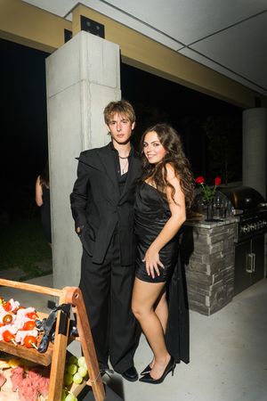Young couple in black formal attire posing outdoors at a nighttime party event.