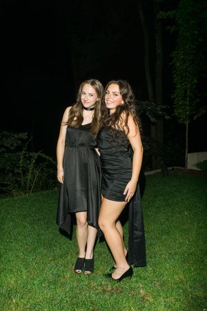 Two young women in black dresses pose together on green grass at night.