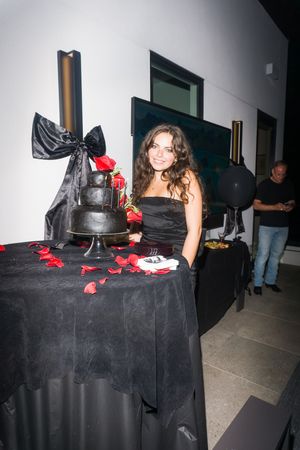 Woman in black strapless dress standing beside elegant black tiered cake with red roses.