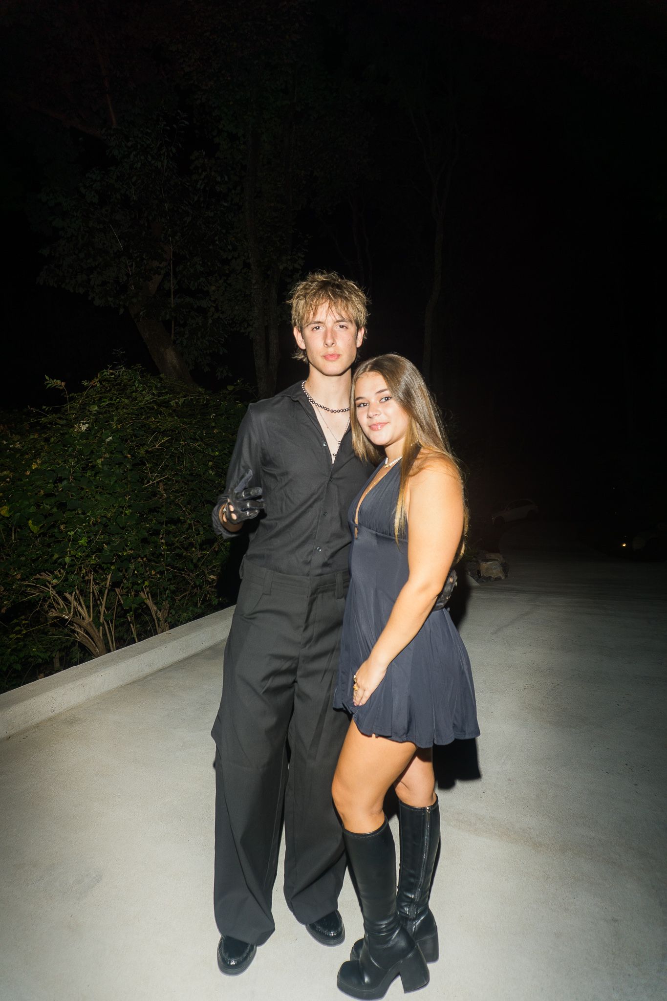 Young couple dressed in dark formal attire posing outdoors at night, woman in mini dress with knee-high boots.