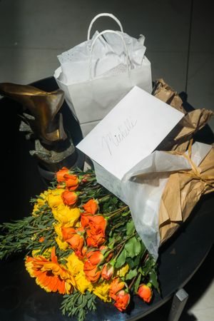 Orange and yellow flowers bouquet with white gift bag, brown paper bag, and a card labeled Michelle on dark table.