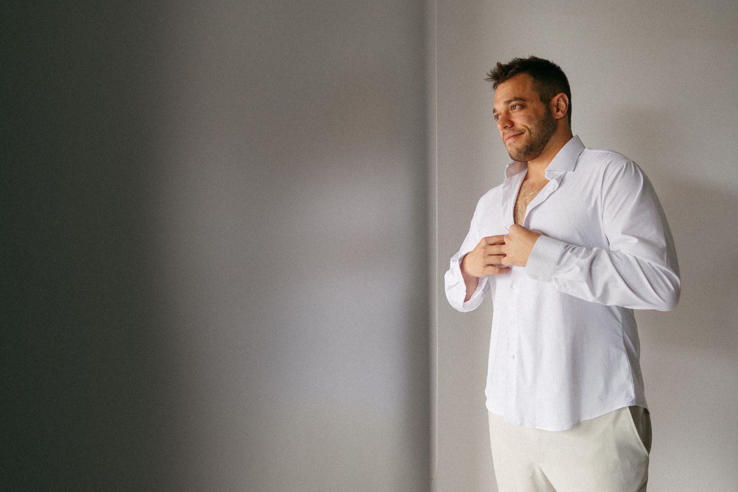 Man in white button-up shirt smiling while getting dressed, standing near a bright window in a modern room.