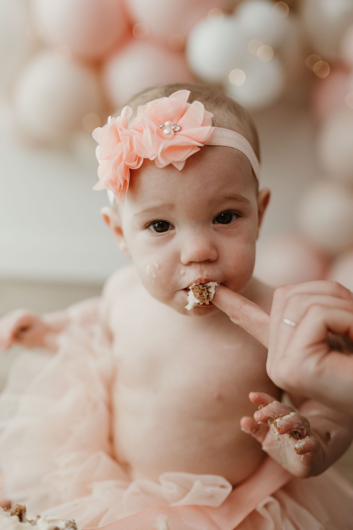 Baby girl in pink tutu and floral headband eating first birthday cake at a smash cake photo session.