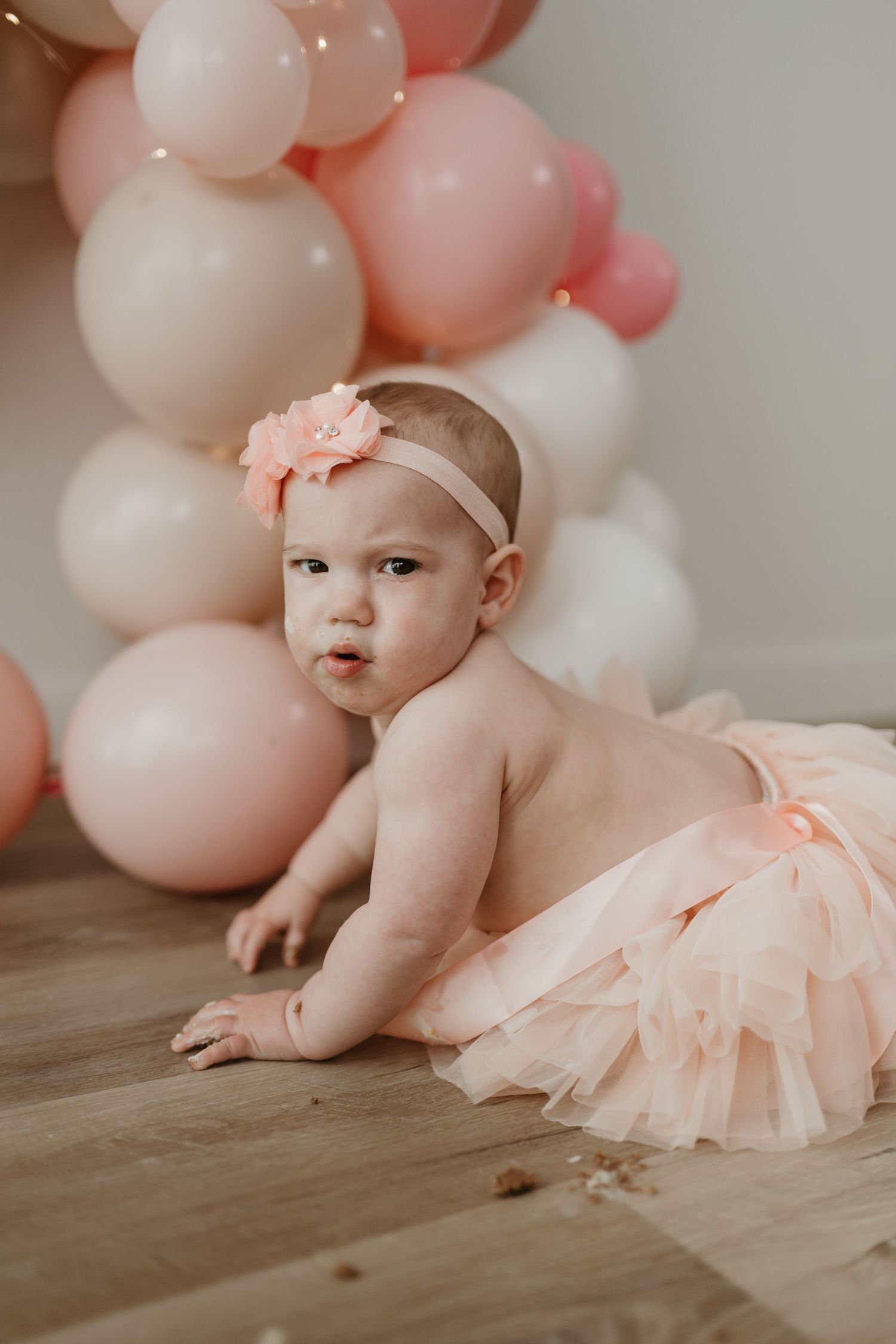 Baby girl in peach tutu skirt and floral headband crawls on wood floor at first birthday party with pink balloon arch.