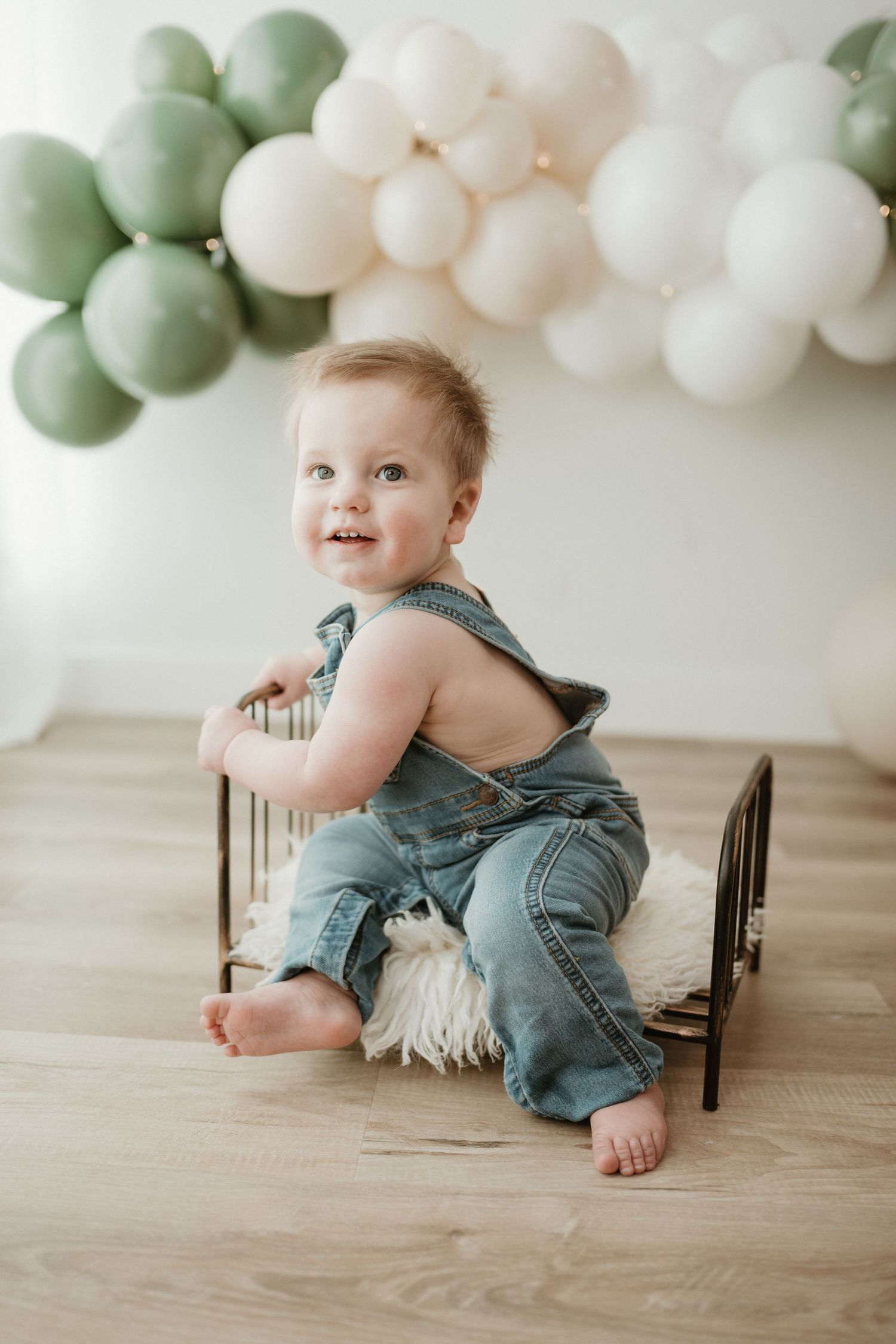 A smiling baby in denim overalls sits on a small bed with green and white balloons in the background. Regina cake smash photographer.