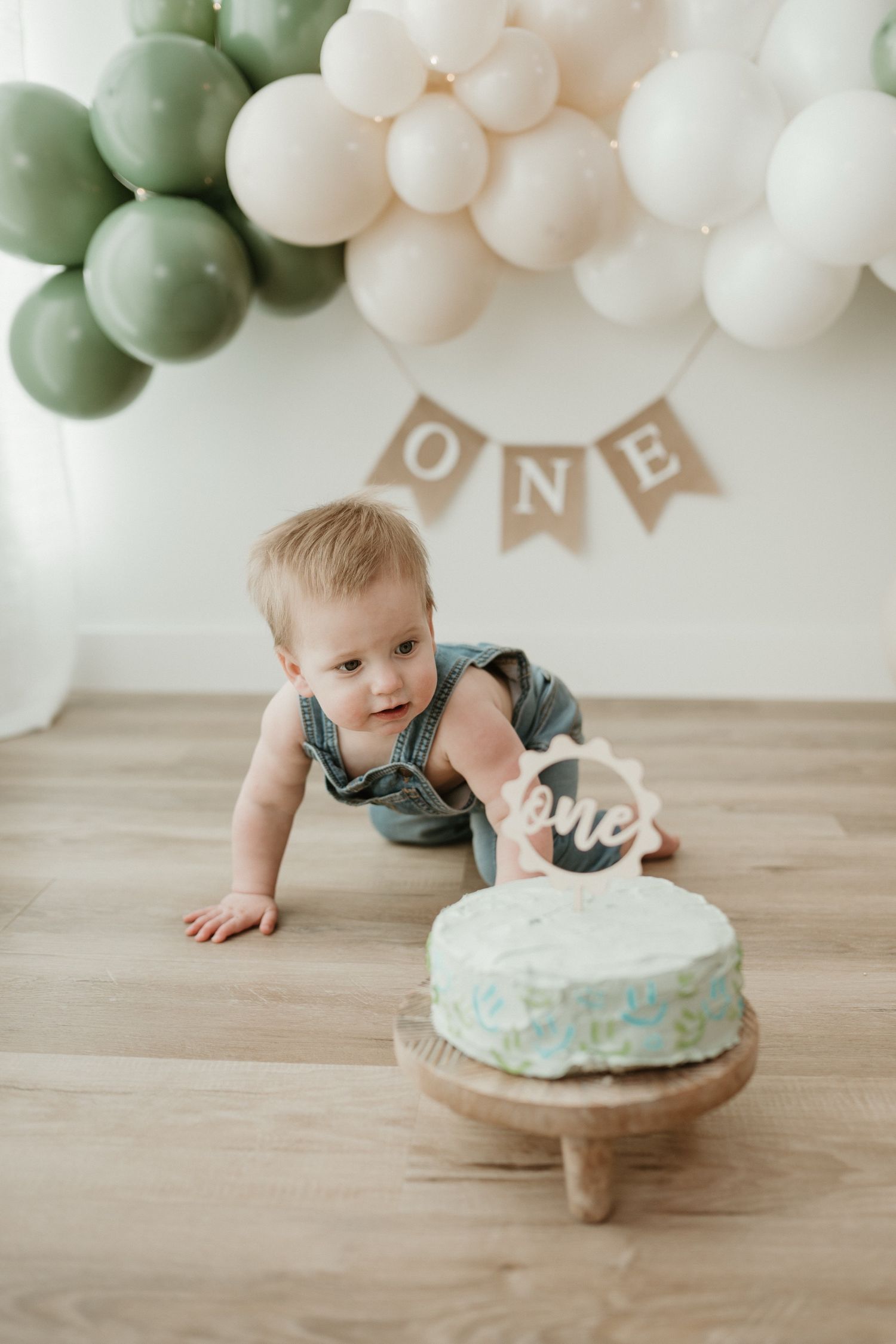 Baby's first birthday cake smash with green and white balloons, ONE banner, and blue cake on wooden stand.
