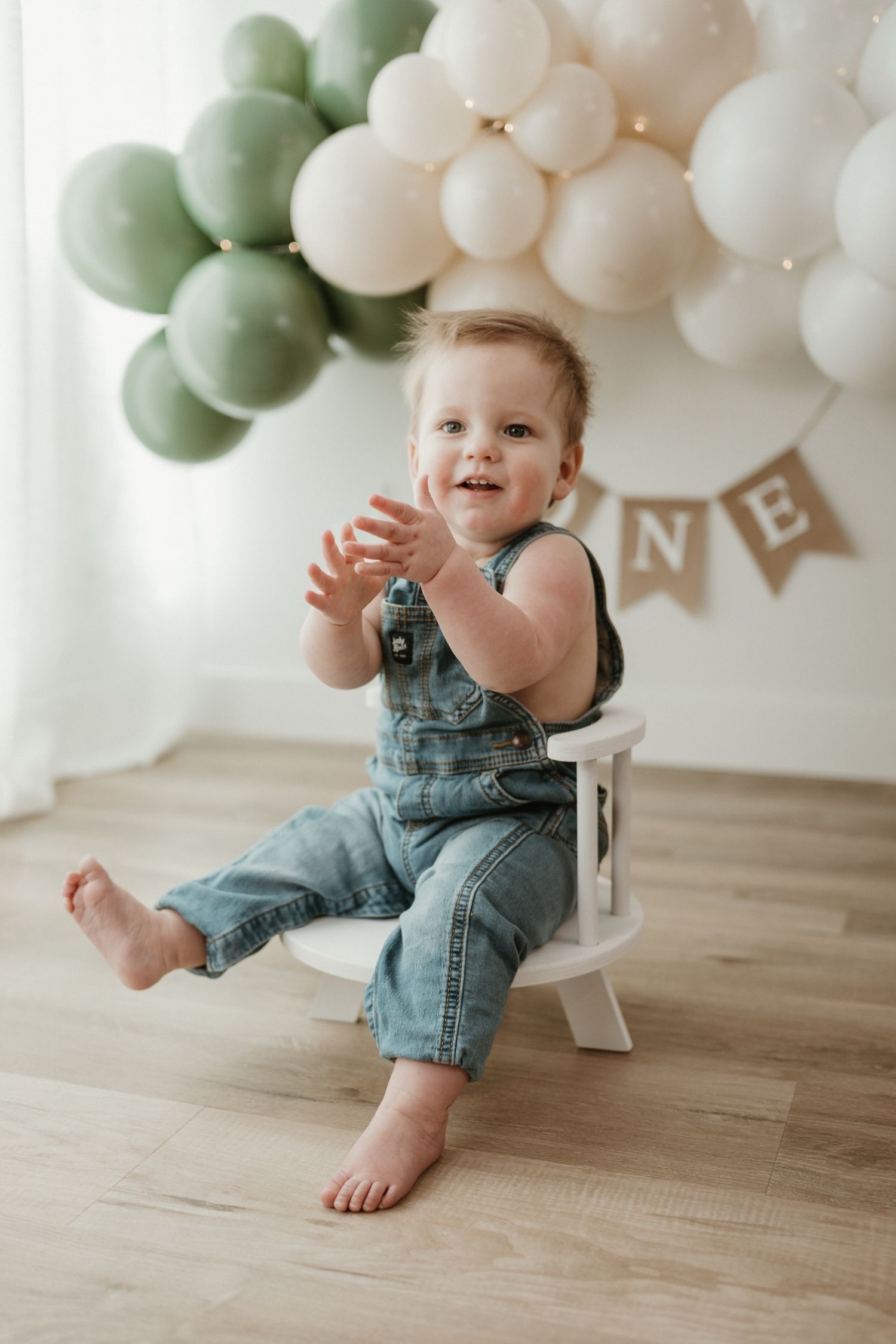 Smiling baby in denim overalls clapping hands, sitting on white chair with green and white balloons for first birthday.