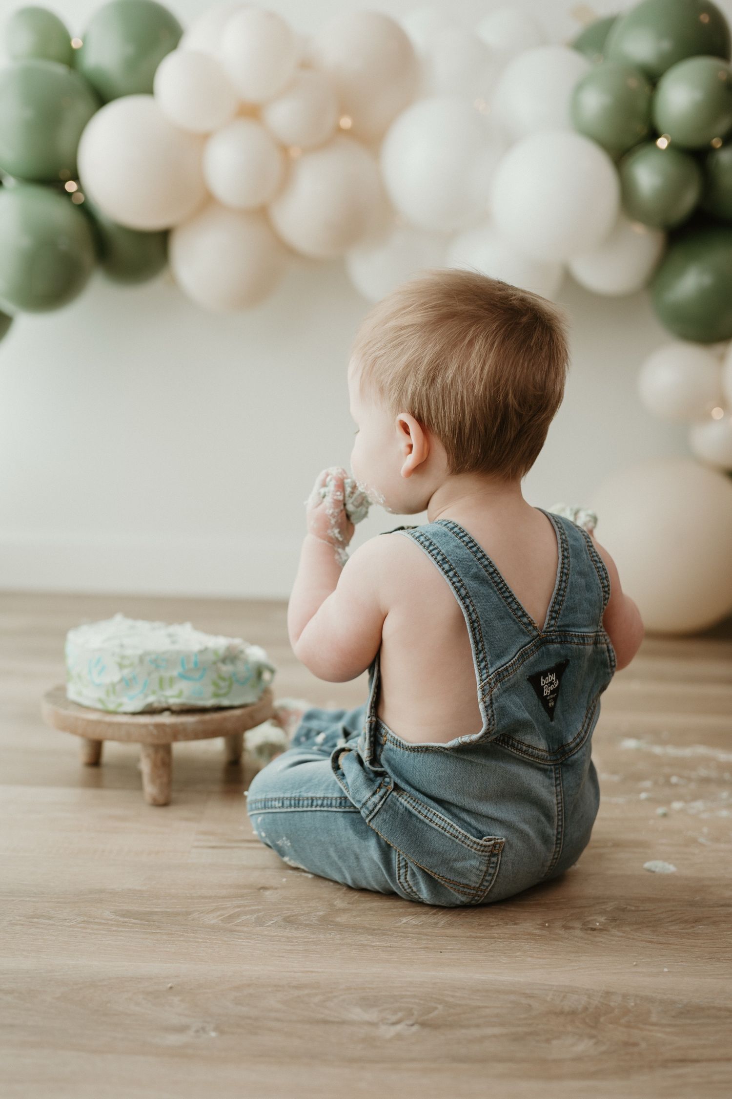 Baby in denim overalls sits on floor smashing first birthday cake, with green and white balloon garland backdrop.