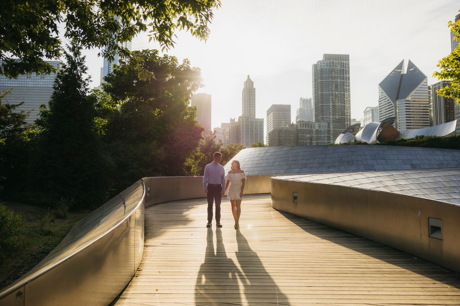Couple walking on BP Pedestrian Bridge with Chicago skyline on background during engagement session at soft sunset light