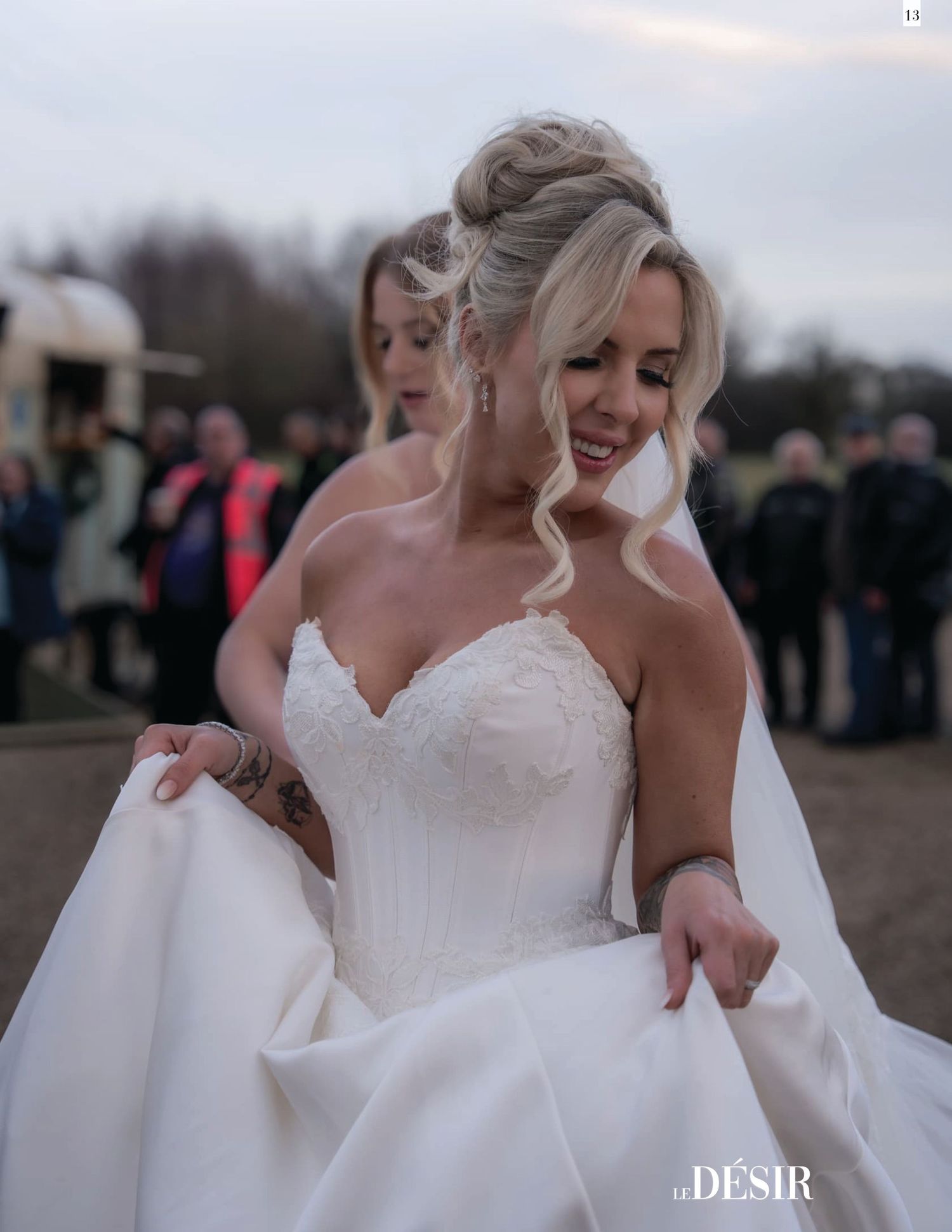 Blonde bride in strapless lace ballgown adjusting her dress outdoors at wedding ceremony.