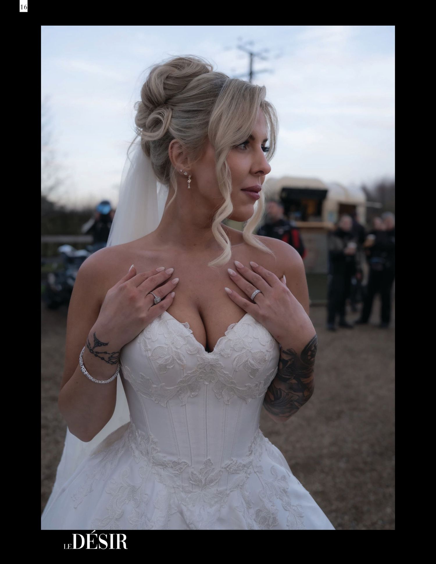 Blonde bride in strapless lace corset wedding gown with veil and tattoos posing outdoors at rustic venue.
