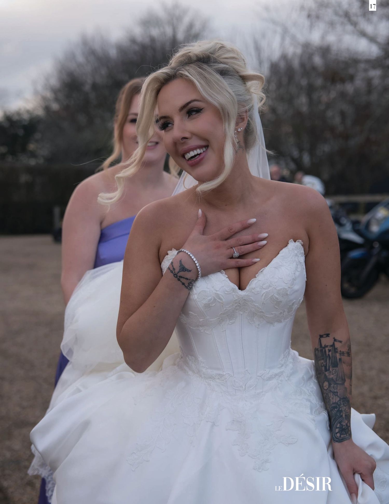 Smiling bride in white strapless lace gown with veil and arm tattoo, bridesmaid in lavender dress behind her outdoors.