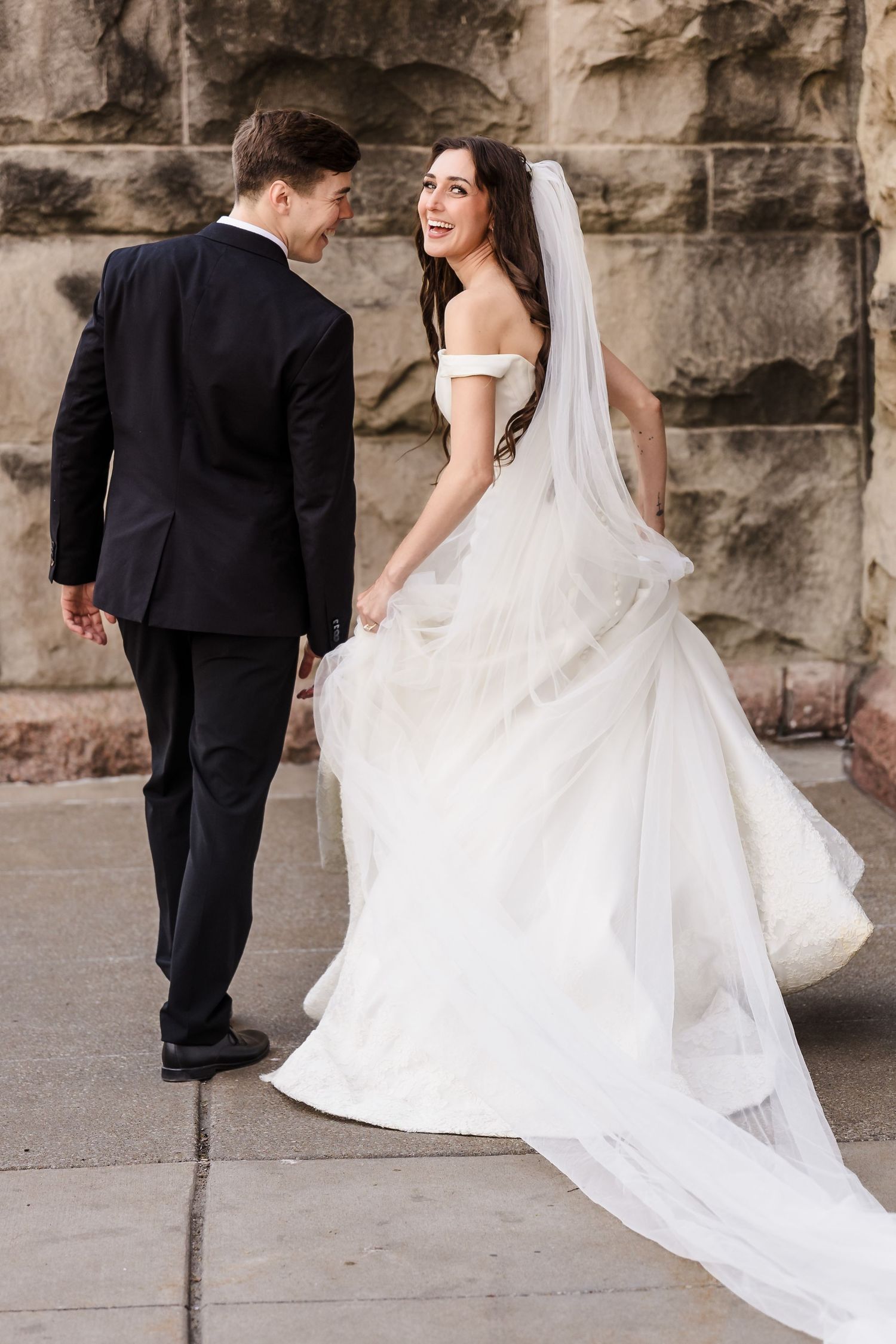 classic wedding portrait of a bride and groom walking together along a historic stone wall, highlighting the elegant flow of her long weddin