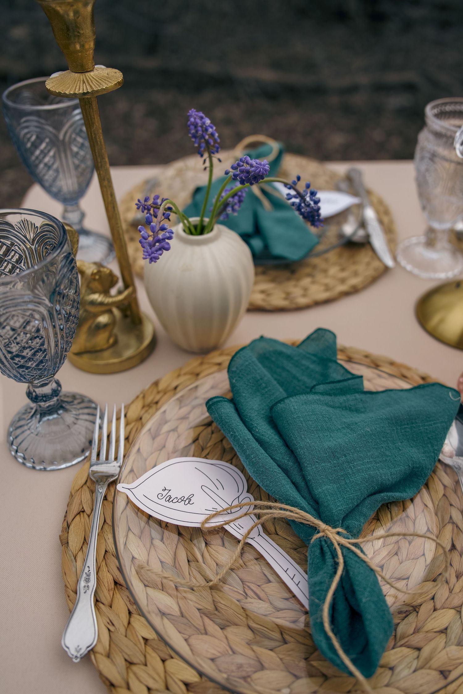 Elegant wedding table setting with teal linen napkin, woven charger, crystal glassware, and purple hyacinth in white vase.