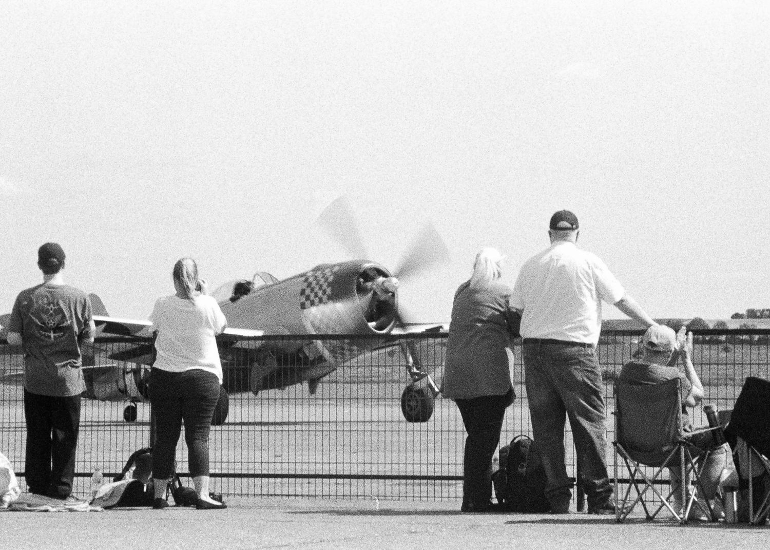 Spectators watch a vintage propeller aircraft taxi on a runway at an outdoor airshow in black and white photo.