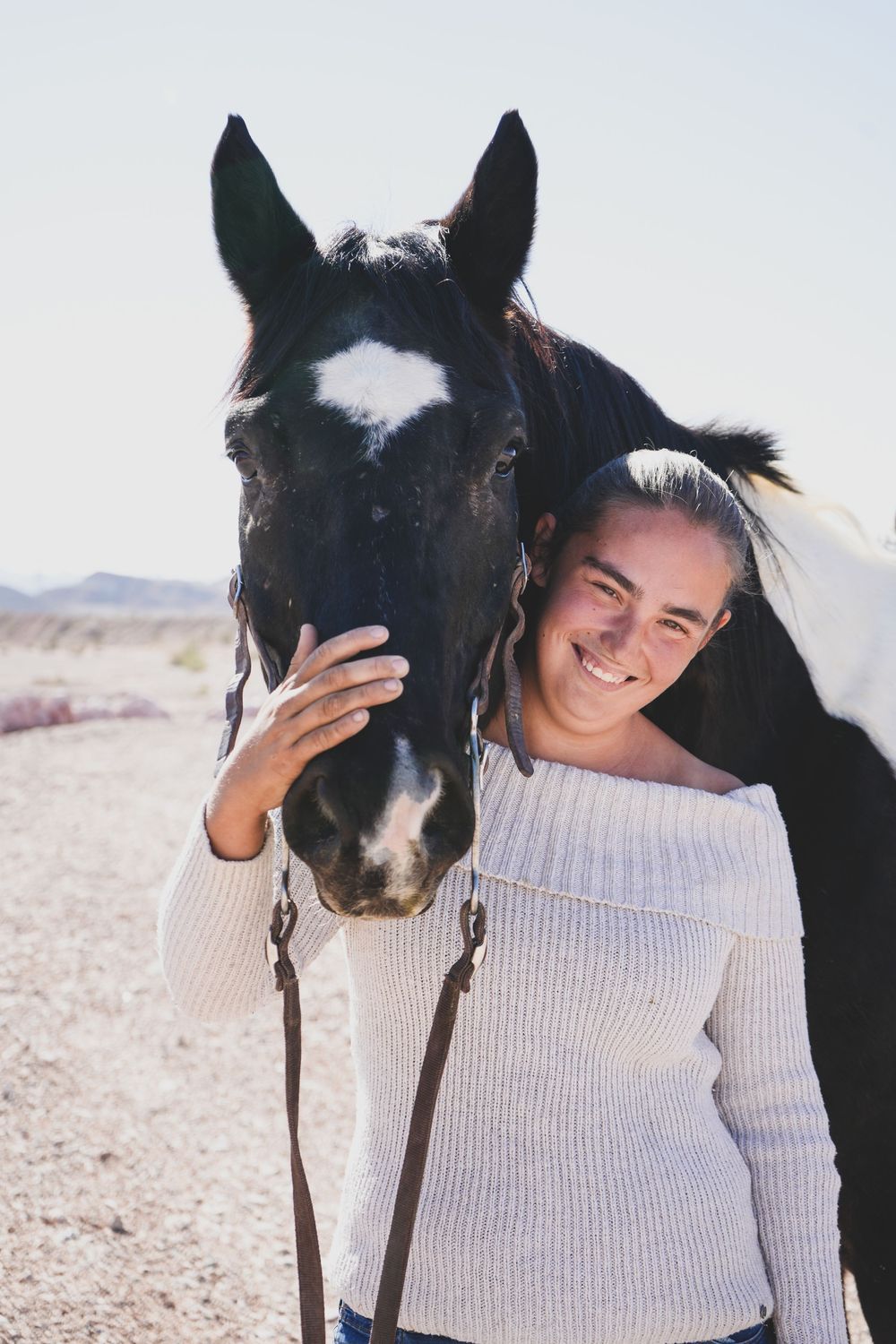 Smiling woman in white sweater posing with a black and white horse outdoors.