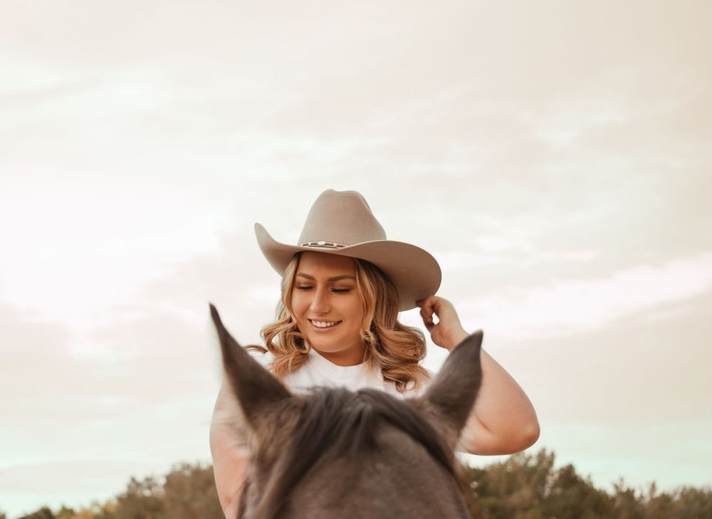 Smiling woman in a cowboy hat sitting on horseback against a warm glowing sky in an outdoor setting.