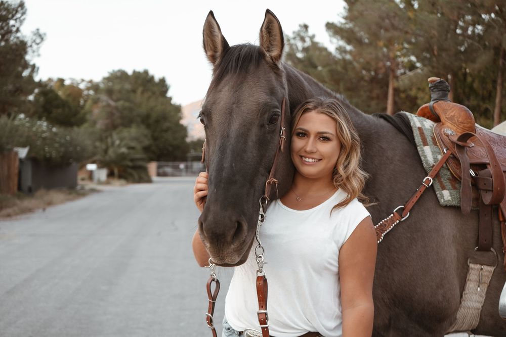 Smiling blonde woman in a white top posing beside a dark brown saddled horse on a tree-lined road.
