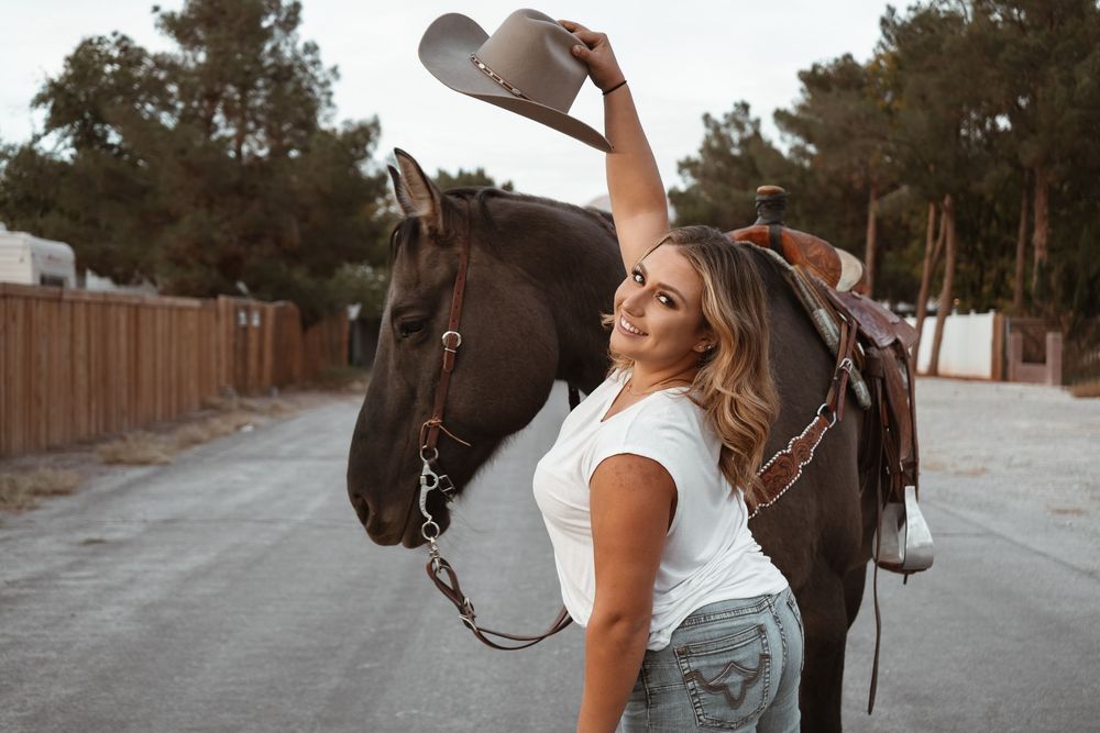 Cheerful woman in jeans raising her cowboy hat while standing beside a dark saddled horse outdoors.