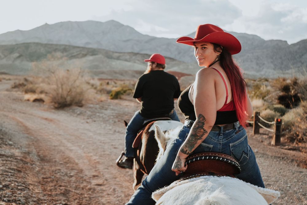 Woman in red cowboy hat rides horse on desert trail, looking back at camera, with mountains in background.