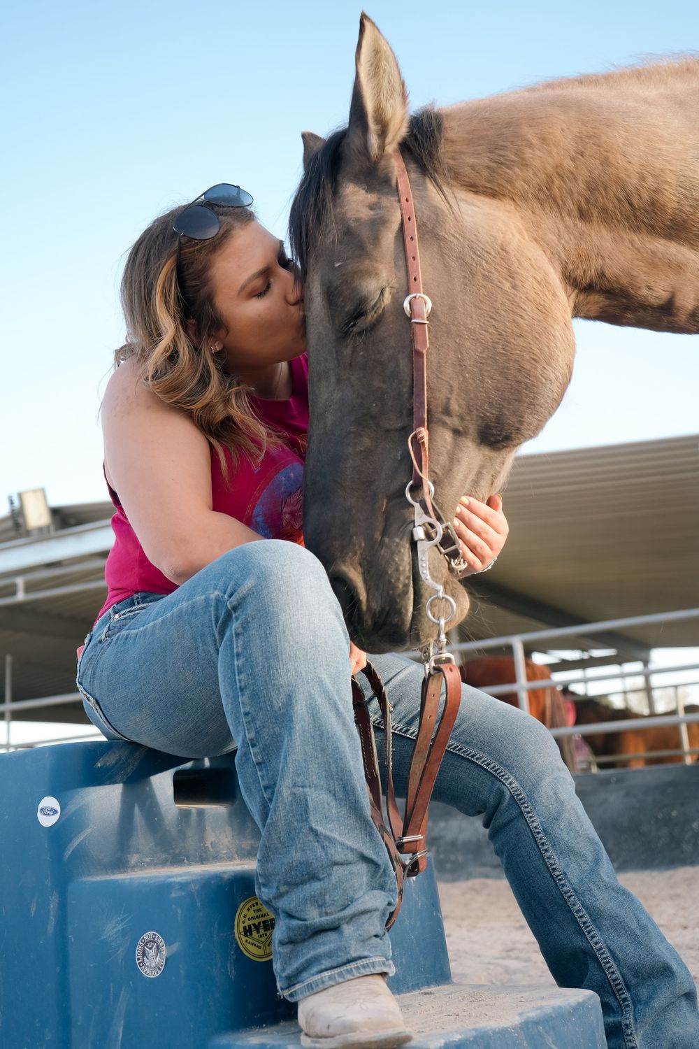 Woman in pink tank top and jeans kissing a brown horse on the nose at a ranch.
