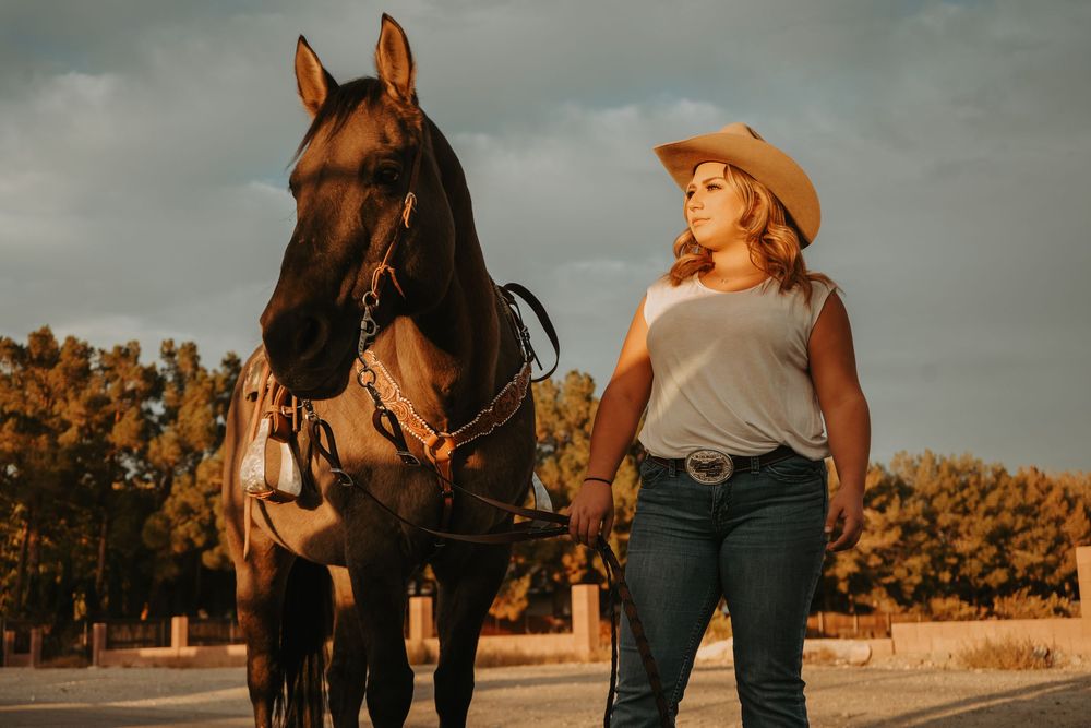 Woman in a cowboy hat and jeans leading a dark horse along a road at golden hour sunset.