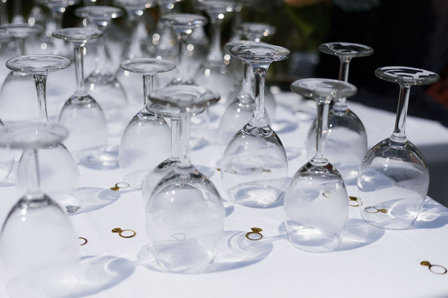 Rows of inverted crystal wine glasses arranged on a white tablecloth with gold ring confetti for a wedding reception.