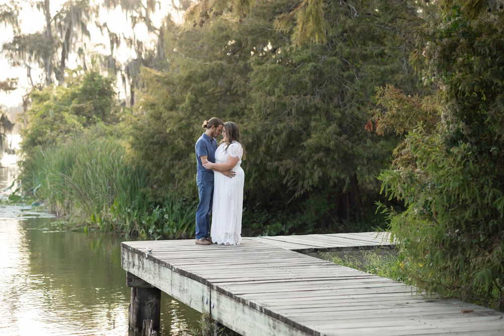 Couple sharing a romantic kiss on a wooden dock surrounded by lush greenery and a serene lake at golden hour.