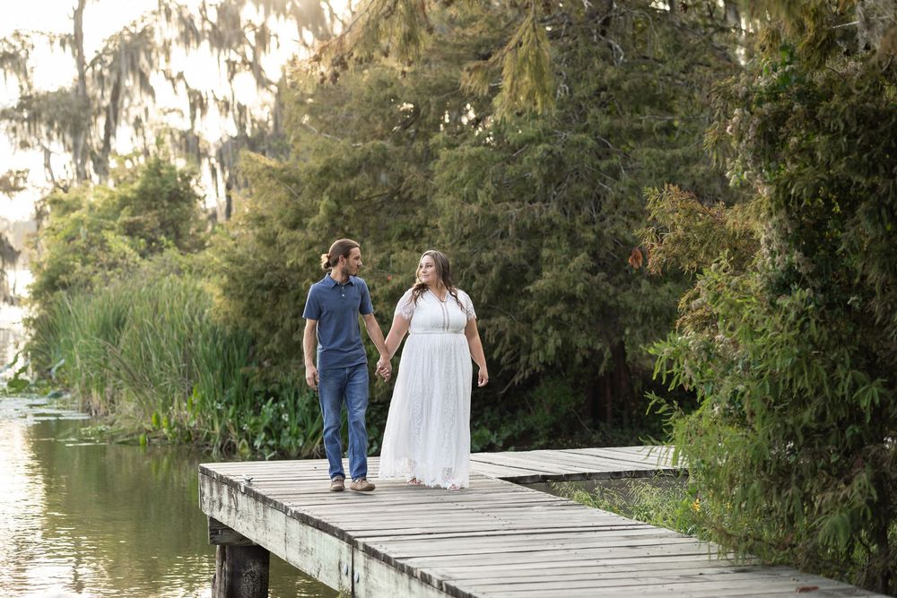 Couple holding hands walking on wooden dock surrounded by lush greenery and water, woman in white dress, maternity photo.