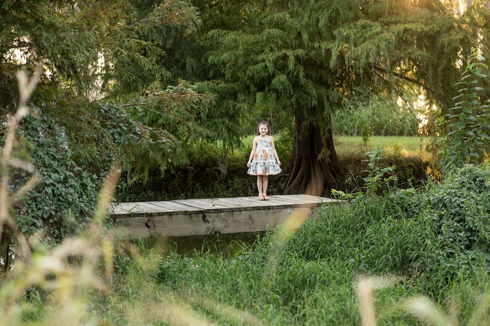 A young girl in a floral dress stands on a stone bridge surrounded by lush green trees and ivy in a park.