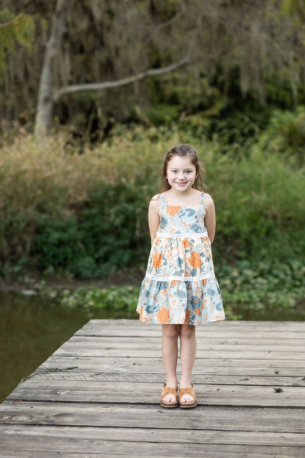 Young girl in floral dress smiling on wooden dock by a lake surrounded by lush greenery.