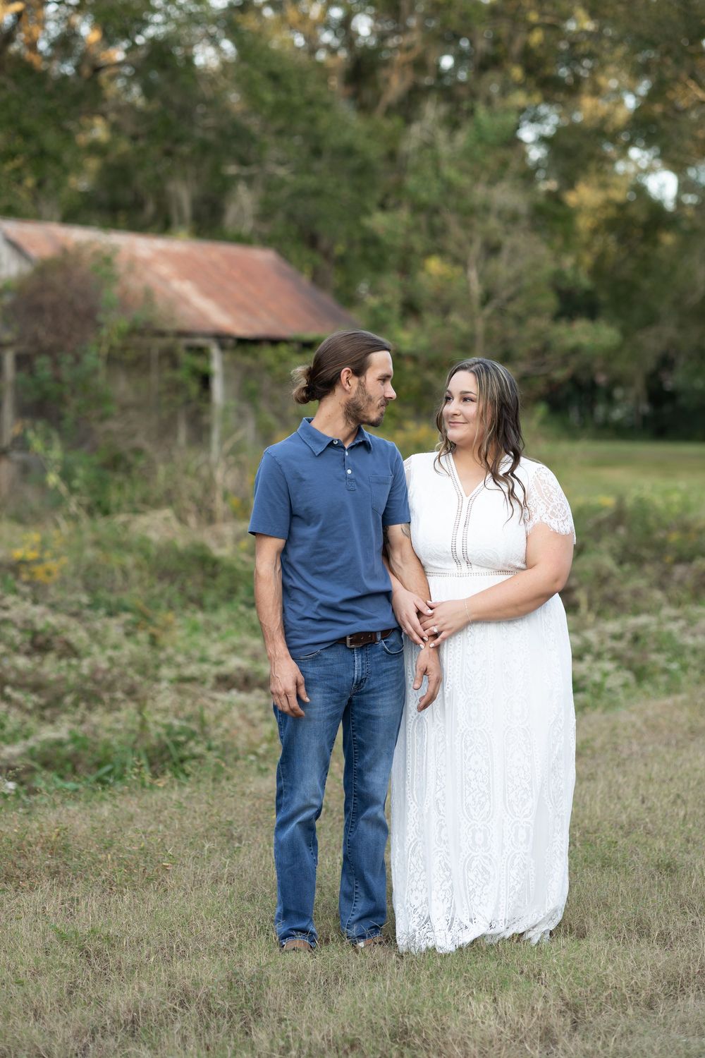 Couple holding hands in a field, woman in white lace dress and man in blue polo and jeans, rustic barn background.