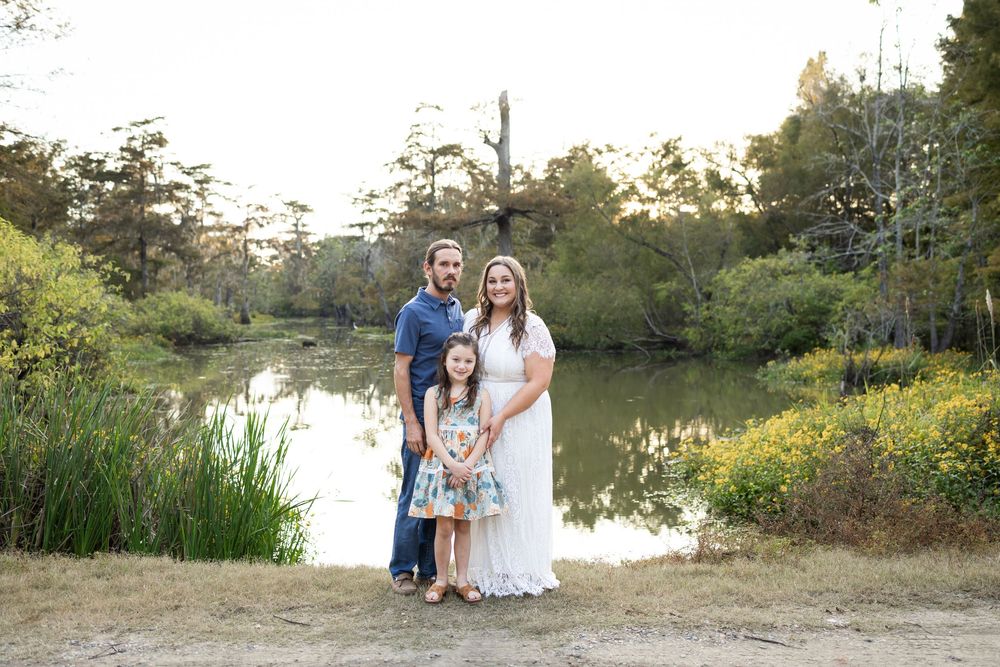 Family of three posing by a serene pond surrounded by lush greenery, with parents and young daughter smiling outdoors.
