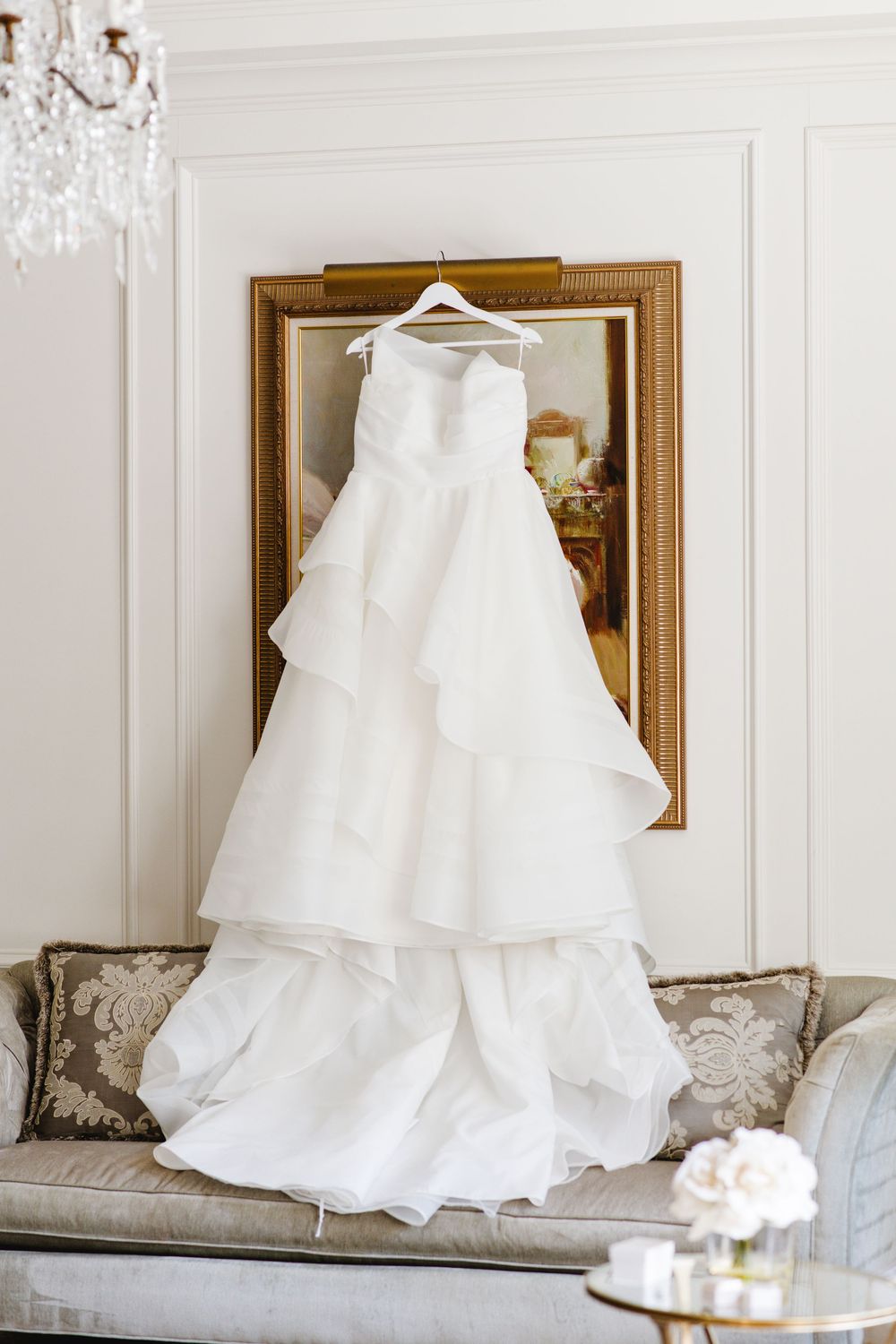 White tiered ball gown wedding dress hanging on hanger in front of gold framed mirror in elegant room.