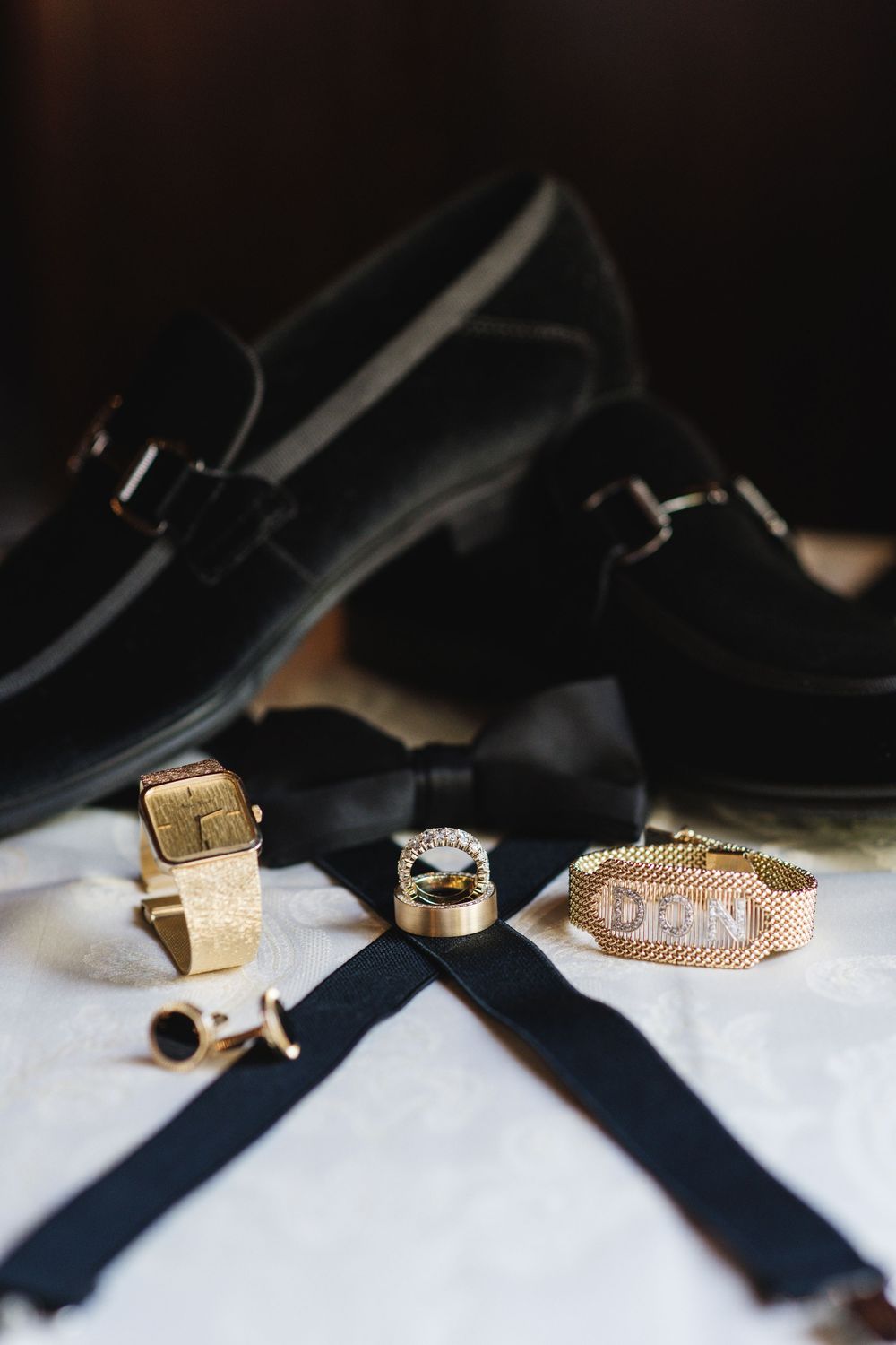 Gold cufflinks, monogram bracelet, diamond ring, black bow tie, suspenders, and loafers arranged as groom wedding accessories.
