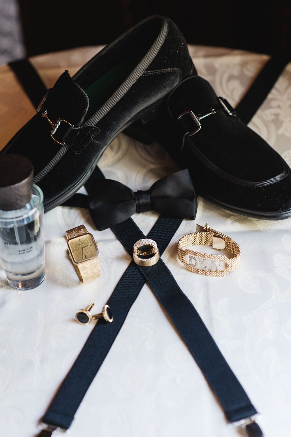 Groom wedding accessories flat lay with black velvet loafers, bow tie, suspenders, gold watch, and cufflinks.